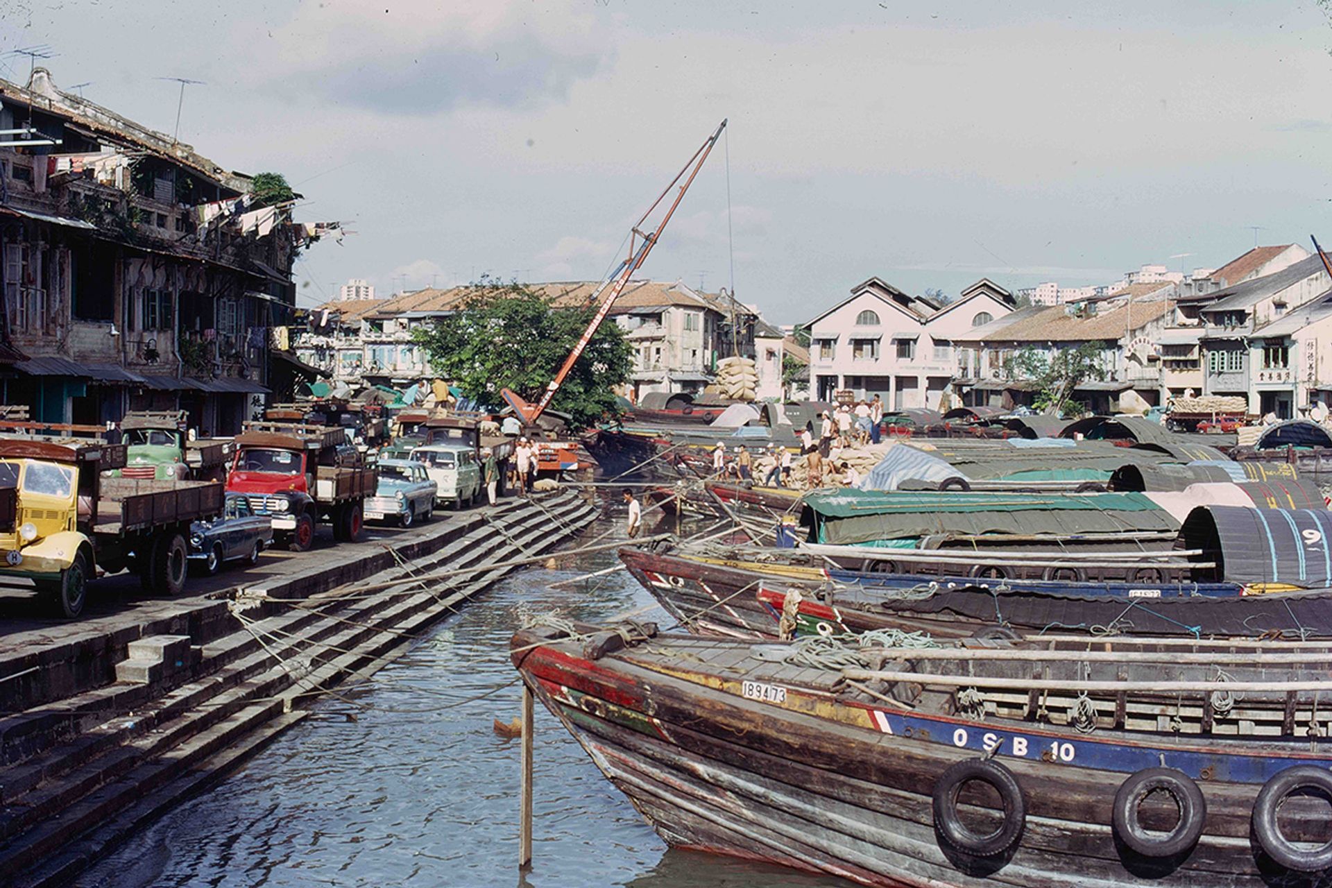 Photographed in the 1960s in Clarke Quay, large boats moored out in the roadstead, and cargo was unloaded into smaller boats known as tongkang or twakow, to be taken up the Singapore River to the riverside godowns or warehouses.