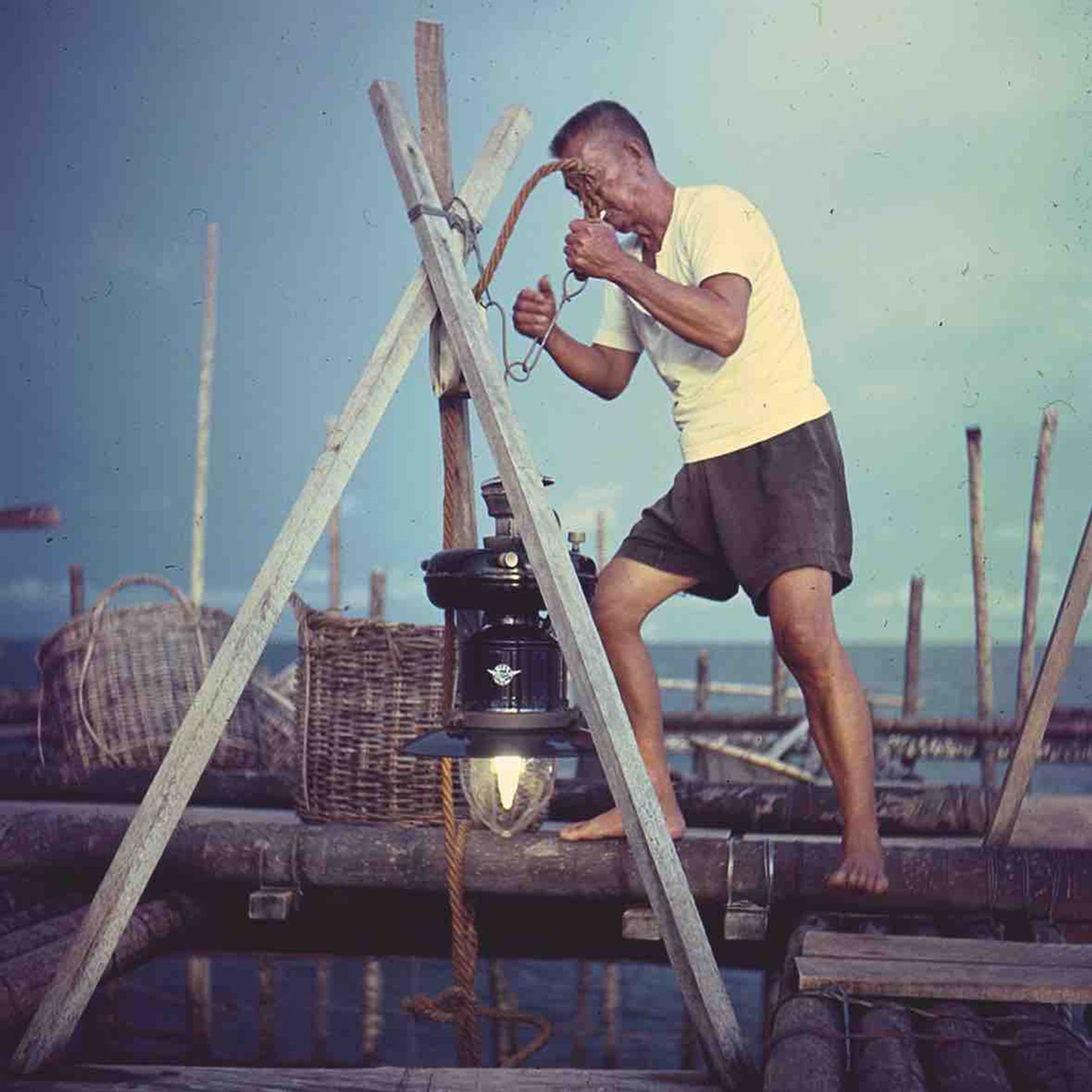 As dusk falls, the kelong is a hive of activity as fishermen prepare themselves for the night's catch. Here you see the powerful kerosene lamps lit, and then lowered just above the water to attract the fish where the net enclosure is located. Photographed in the late 1960s at a kelong off Tuas.