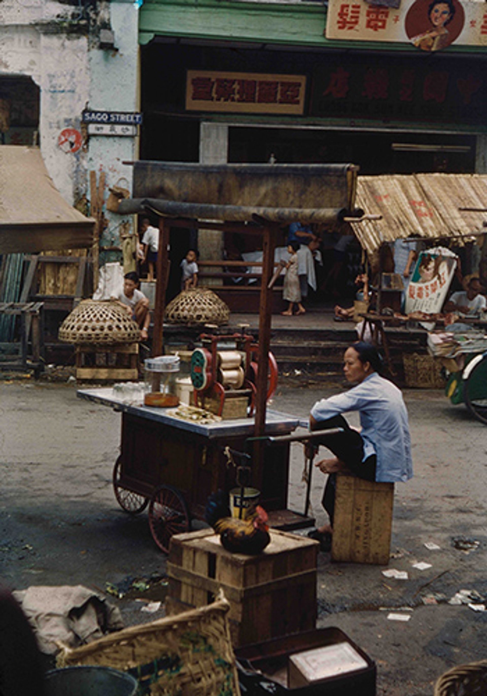 A hawker waiting for customers while resting on a wooden crate beside her hand-operated sugar cane juicer in Sago Street in 1954.