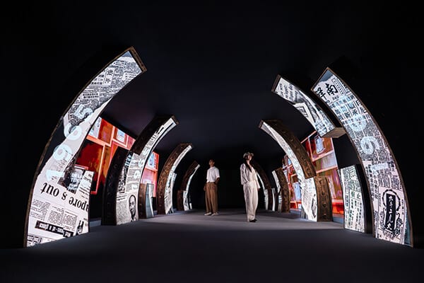 Immersive exhibit with archways projecting historical newspaper content; two people stand inside.