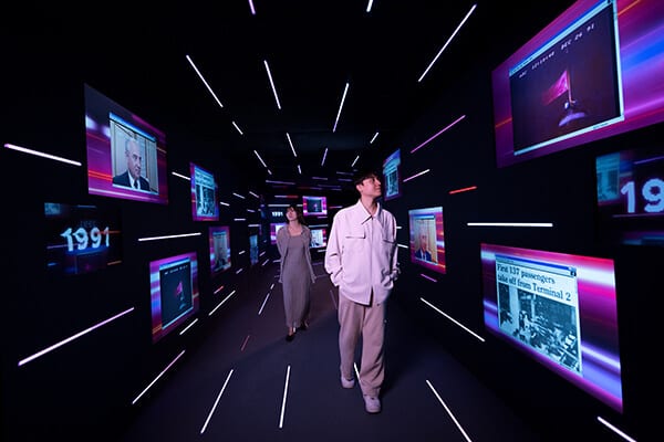 Two people exploring a dark, neon-lit immersive exhibit with historical images and dates on screens.