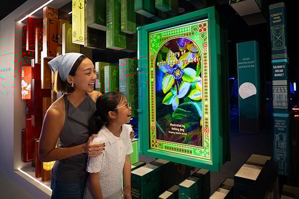 A woman and girl smiling at a glowing digital book cover in a library exhibition.