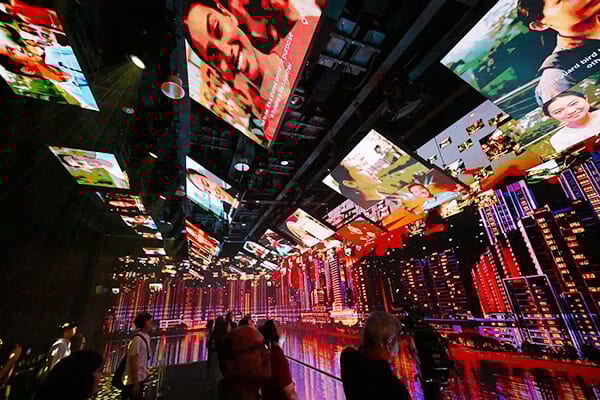 Immersive digital exhibit with multiple screens showing faces and a vibrant city, viewed by people.