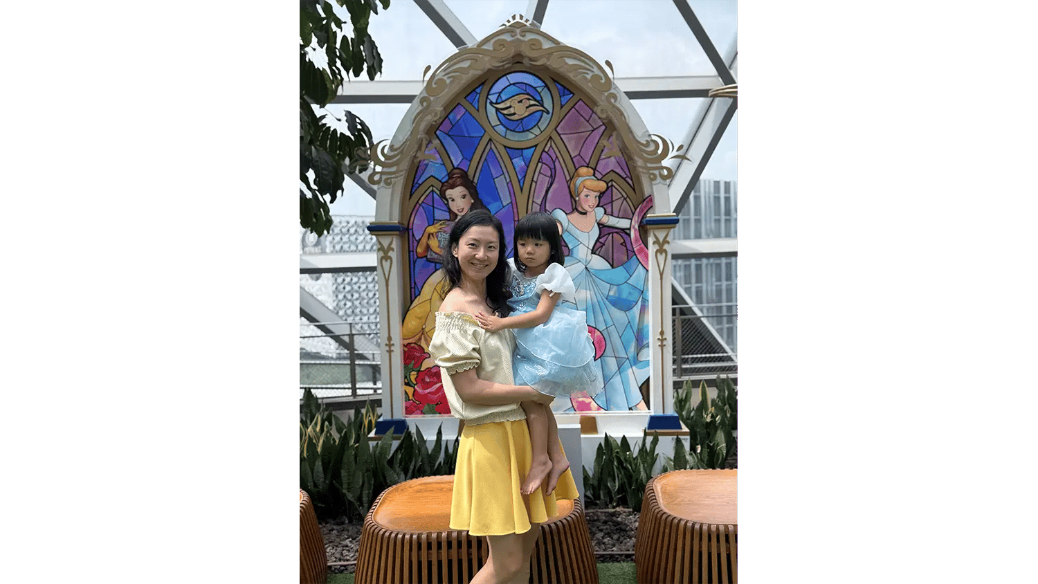 A woman holds a young girl in front of a colorful stained-glass-style Disney Princess display.