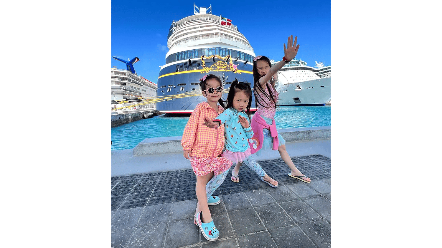 Three young girls pose playfully in front of the Disney Wish cruise ship and other cruise ships.