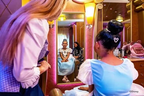 A girl in a blue princess dress smiles at her reflection as a stylist works on her hair.