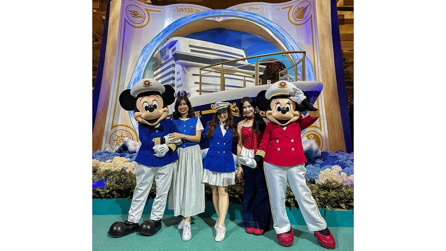 Mickey and Minnie Mouse with three women in nautical outfits pose in front of a Disney Cruise Line ship backdrop.