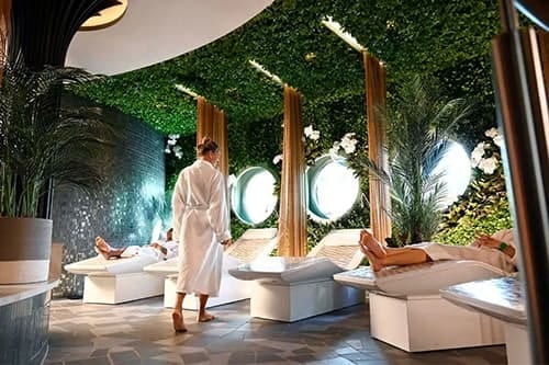 Relaxing spa lounge with green walls, people in robes, and round windows.