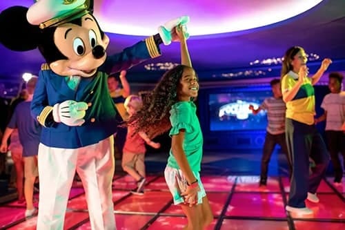Mickey Mouse in a captain's uniform dances with a young girl on a light-up disco floor.