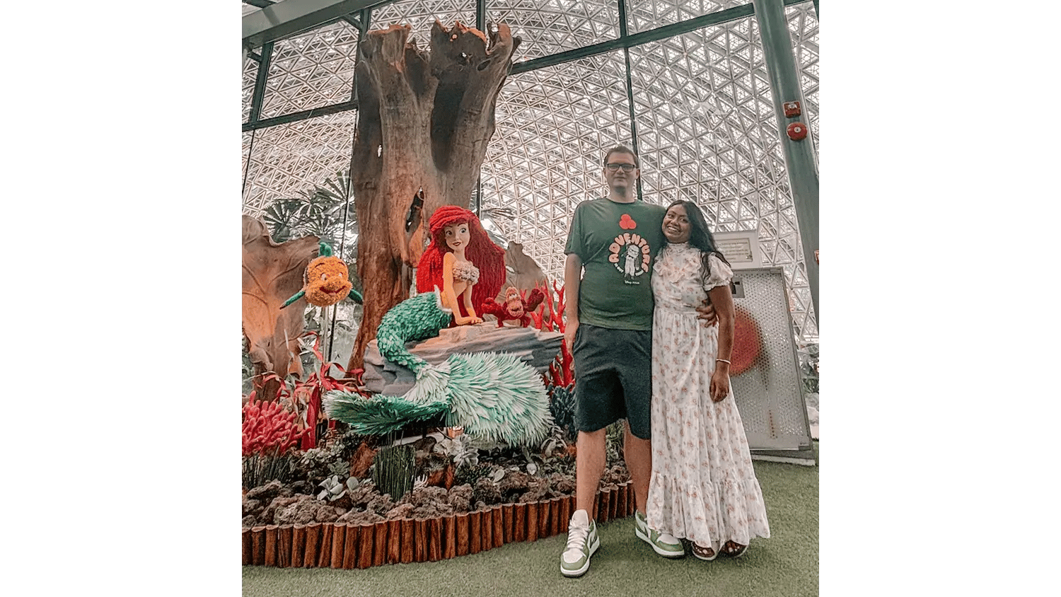 A couple posing happily next to Little Mermaid, Flounder, and Sebastian statues in a large indoor garden.