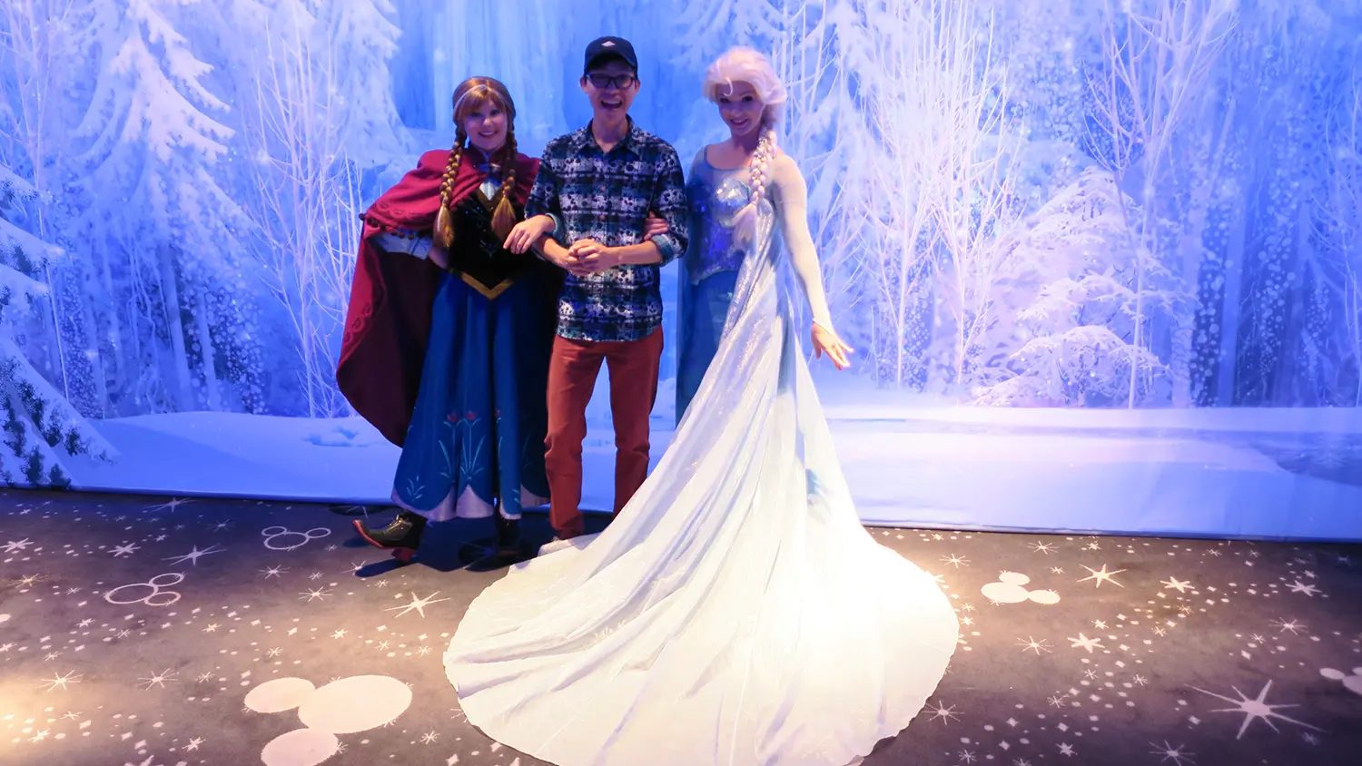 A man poses with Anna and Elsa from Frozen in a winter wonderland backdrop with Mickey Mouse symbols on the floor.