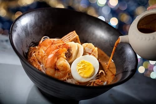 Black bowl of noodles with shrimp, egg, and broth pouring from a teapot.