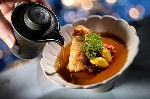 A hand pours dark sauce from a black ceramic pitcher onto a garnished dish in a white bowl.