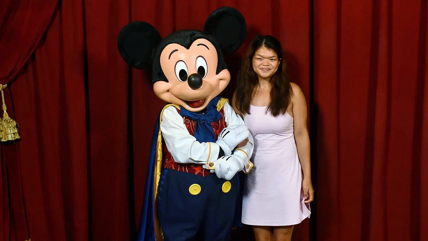 A woman in a pink dress smiles next to Mickey Mouse in a prince costume, against a red curtain backdrop.