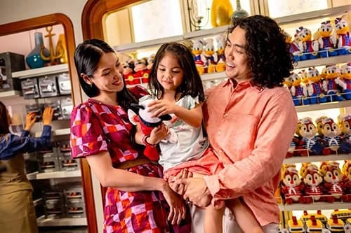 Happy family in a store, child holding a plush toy, surrounded by shelves of Disney character dolls.