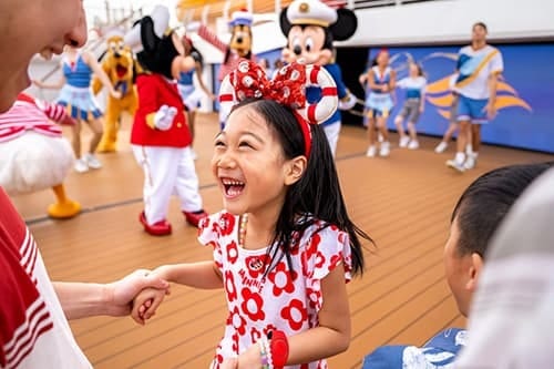 Joyful girl in Minnie headband holds hand; Disney characters dance on deck.