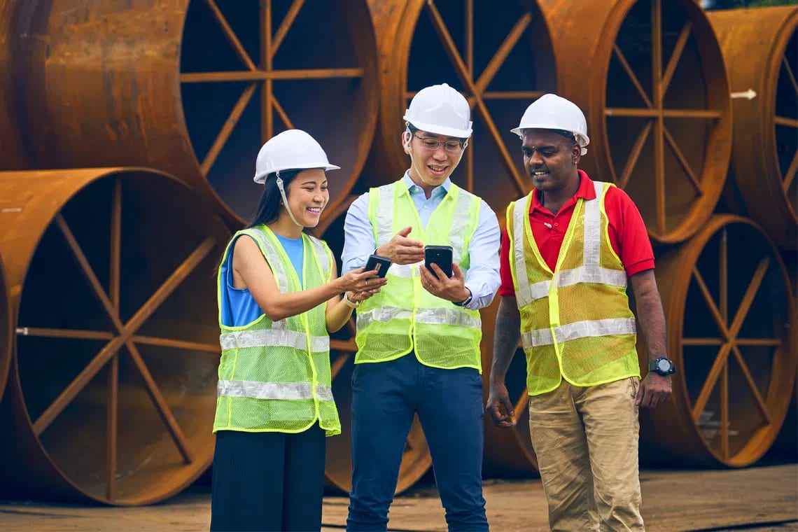 Three construction workers in hard hats and safety vests review information on smartphones at a worksite.