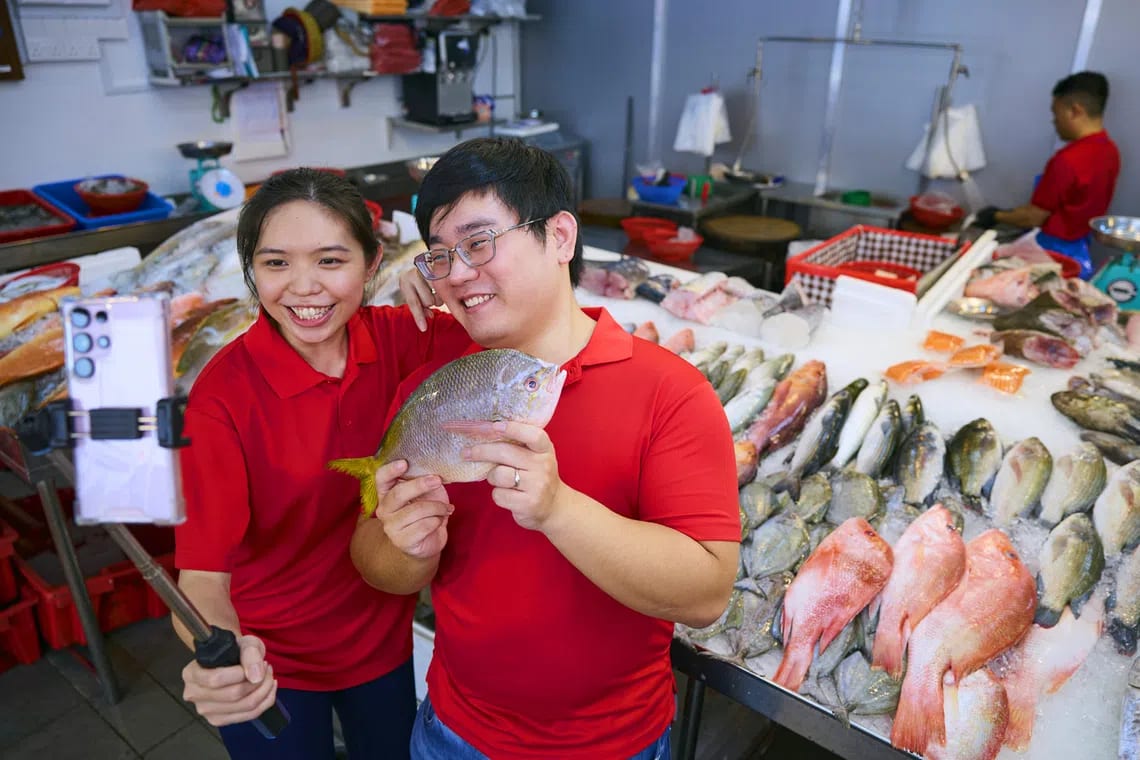 Two smiling individuals in red shirts take a selfie with a fish in a bustling seafood market.