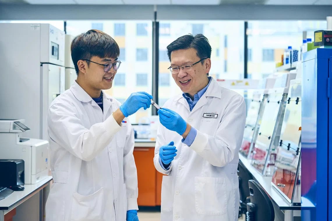 Two scientists in lab coats and gloves smiling and examining a sample tube in a modern laboratory.