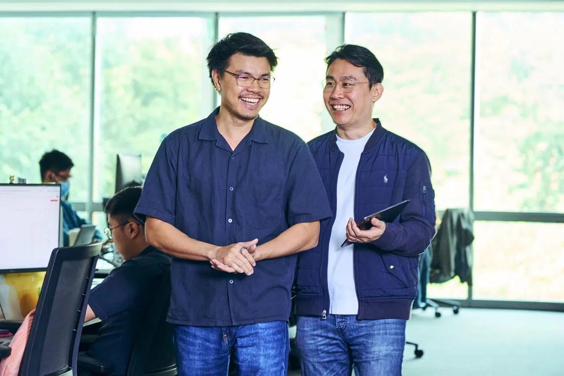 Two smiling men in an office, one holding a tablet, with colleagues in the background.