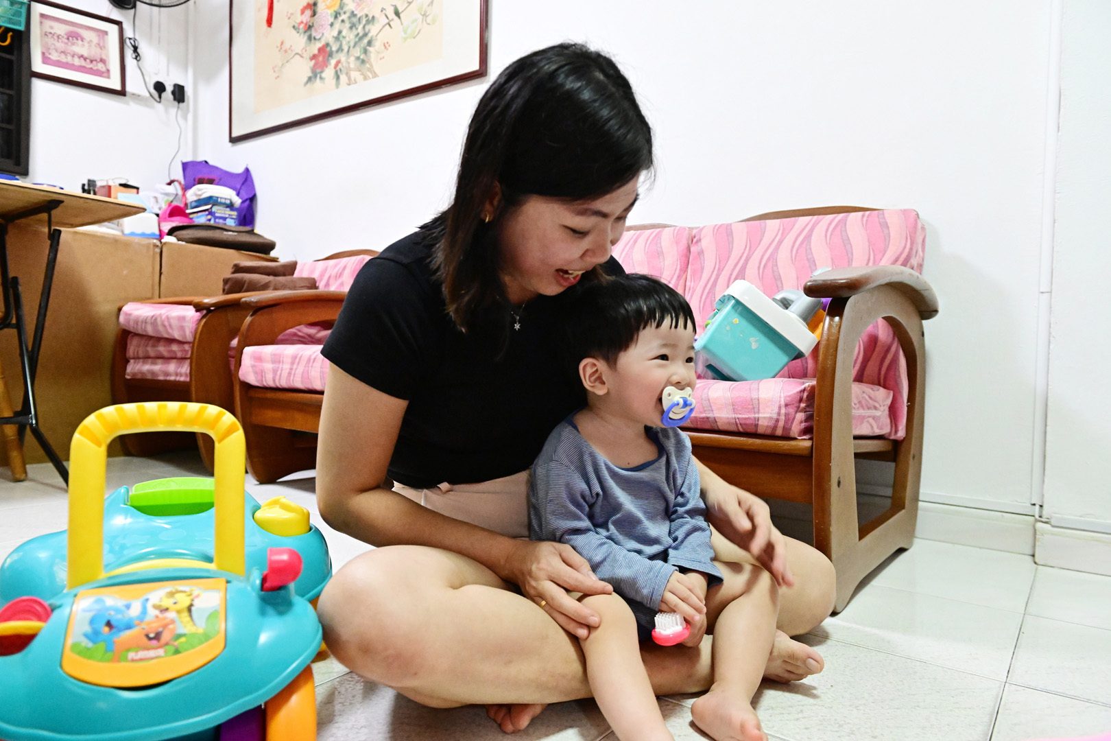 A smiling woman and a young child with a pacifier sit on the floor, looking to the right.