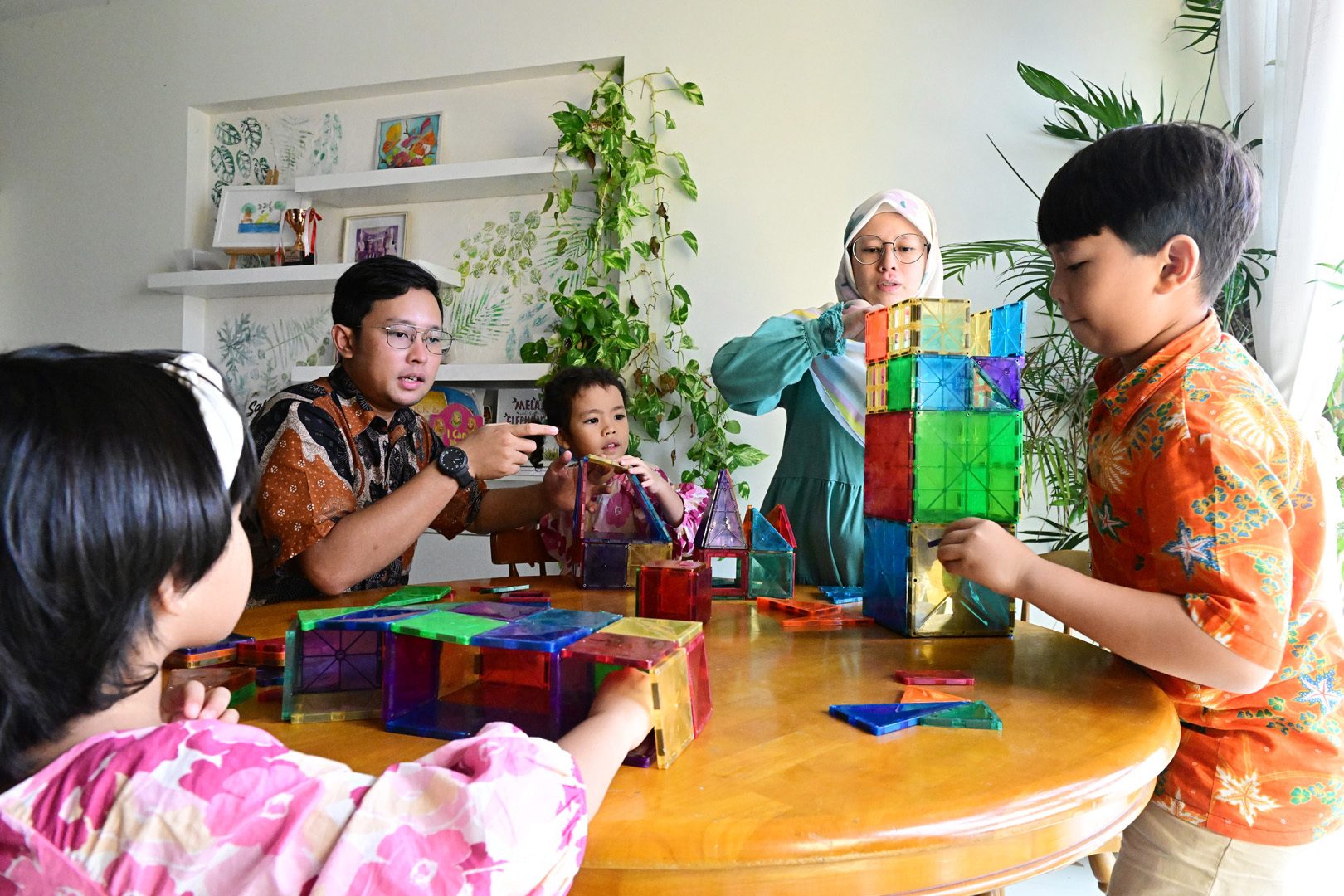 A family, including two adults and three children, plays with colorful magnetic building blocks around a wooden table.