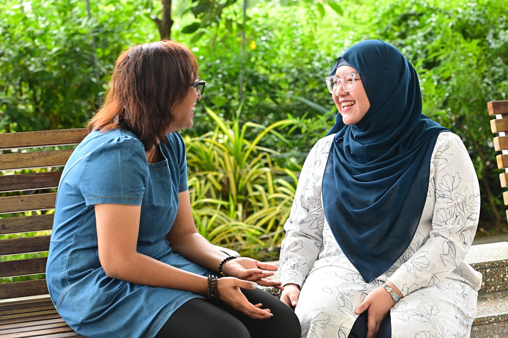 Two women, one in a hijab, happily converse on a bench in a lush green outdoor setting.