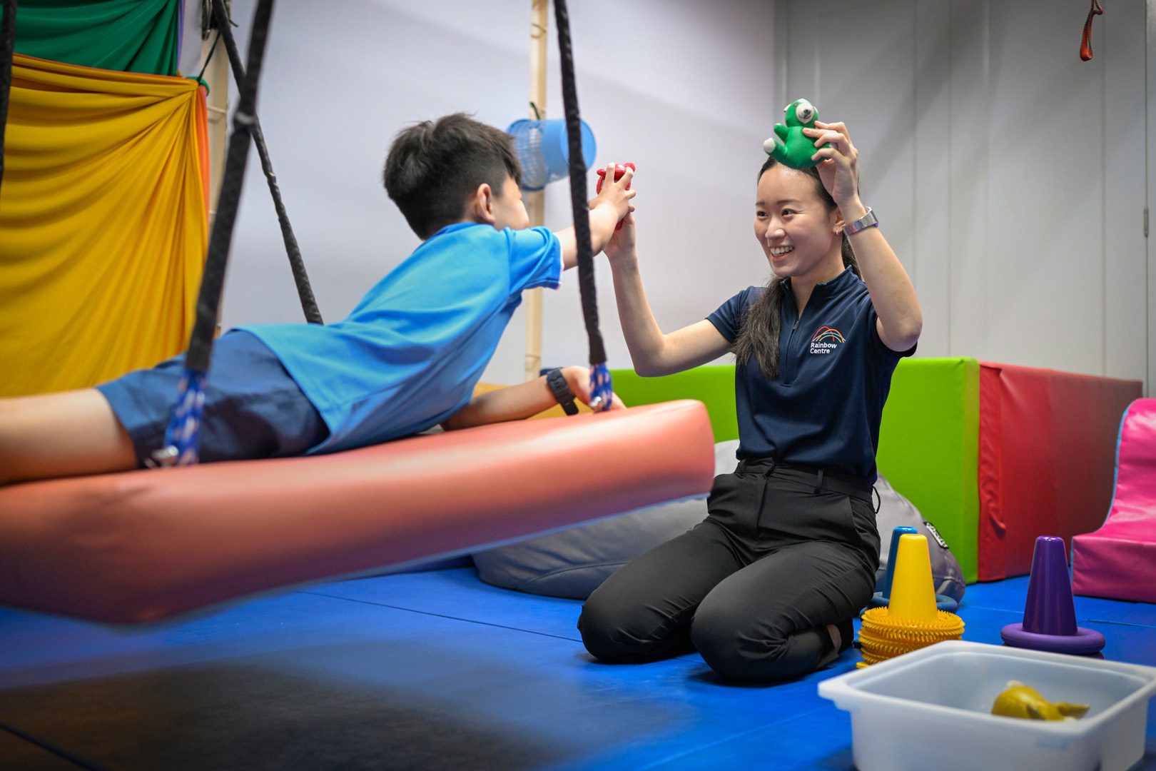 A therapist smiles while playing with a boy on a swing in a colorful therapy room.