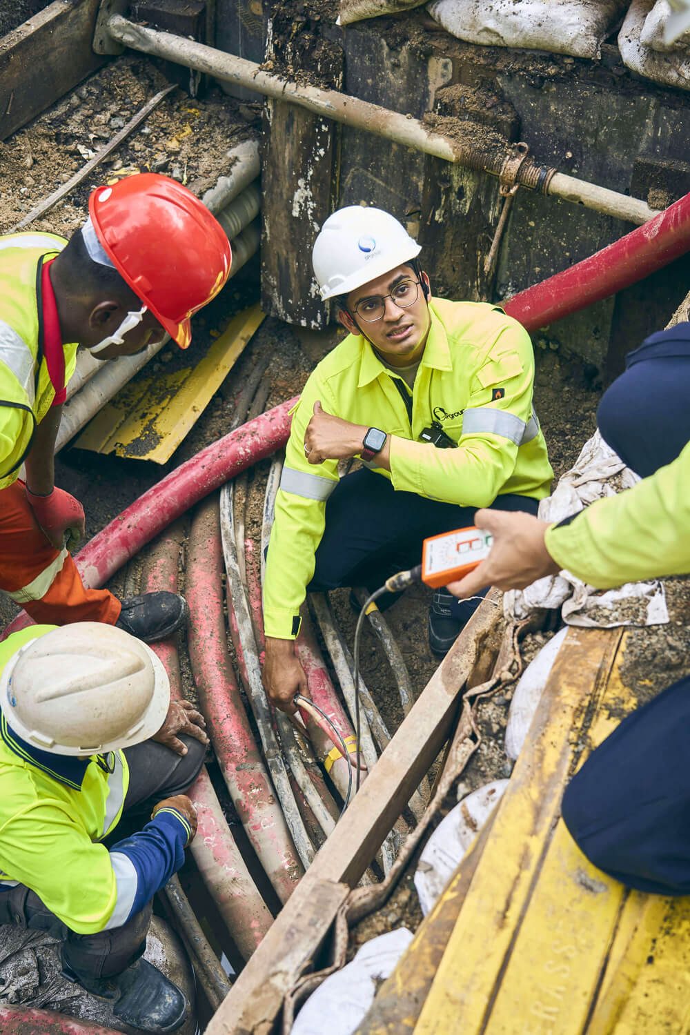 Construction workers in hard hats and hi-vis vests inspect underground cables in a trench.