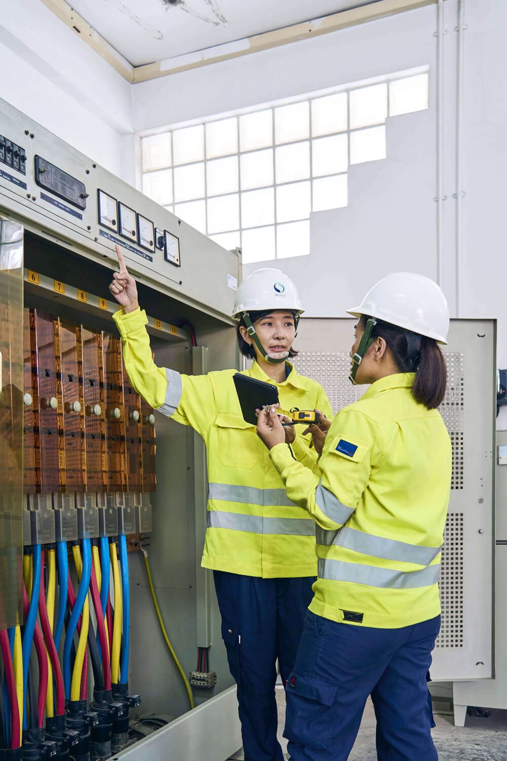 Two engineers in safety gear inspect an electrical panel, discussing measurements and operations.