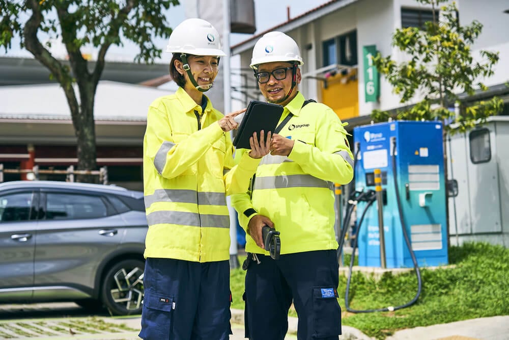 Two smiling technicians in hard hats and yellow vests review data on a tablet at an EV charging station.