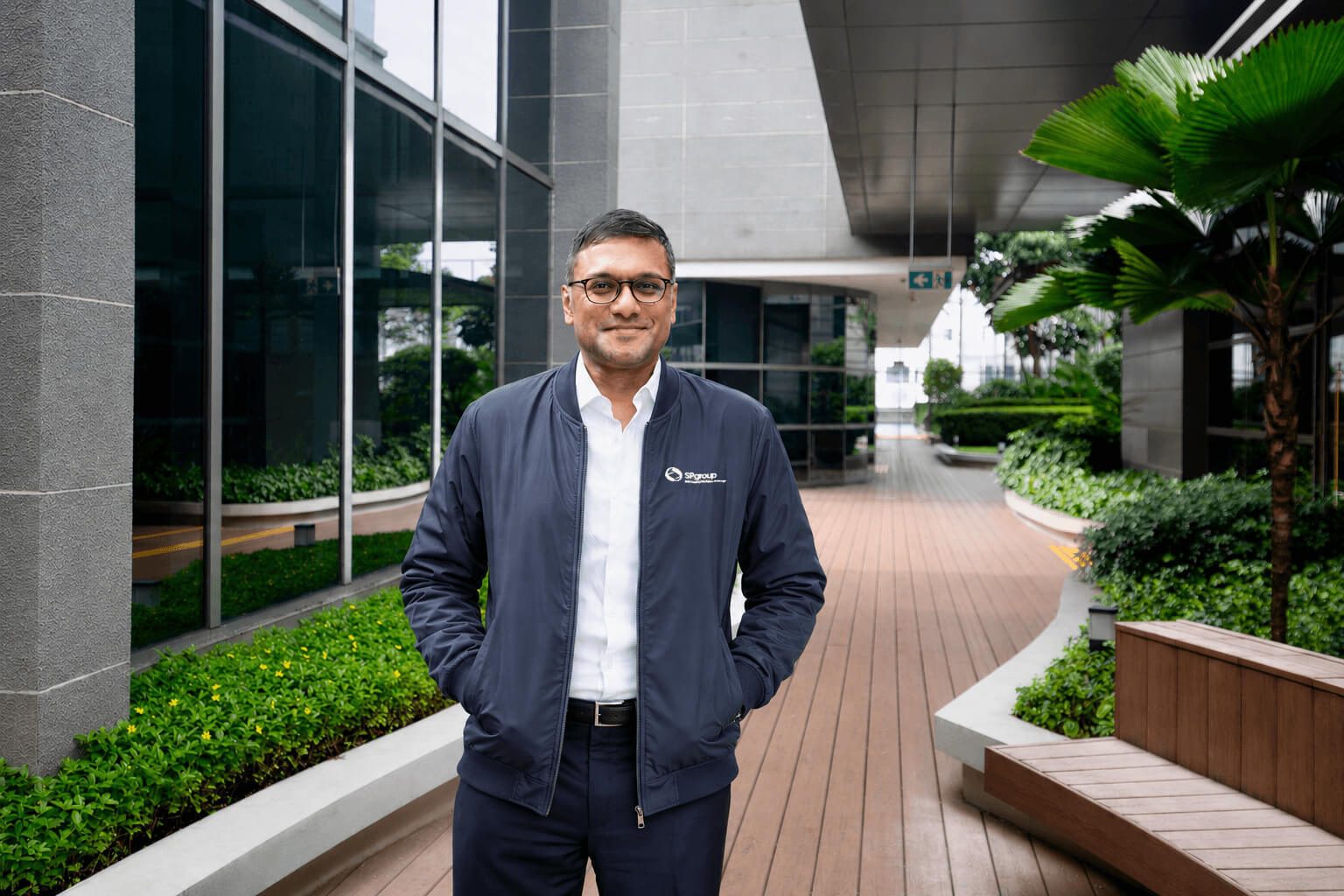 A man in a navy blue jacket and glasses stands outdoors in a modern building complex, looking at the camera.