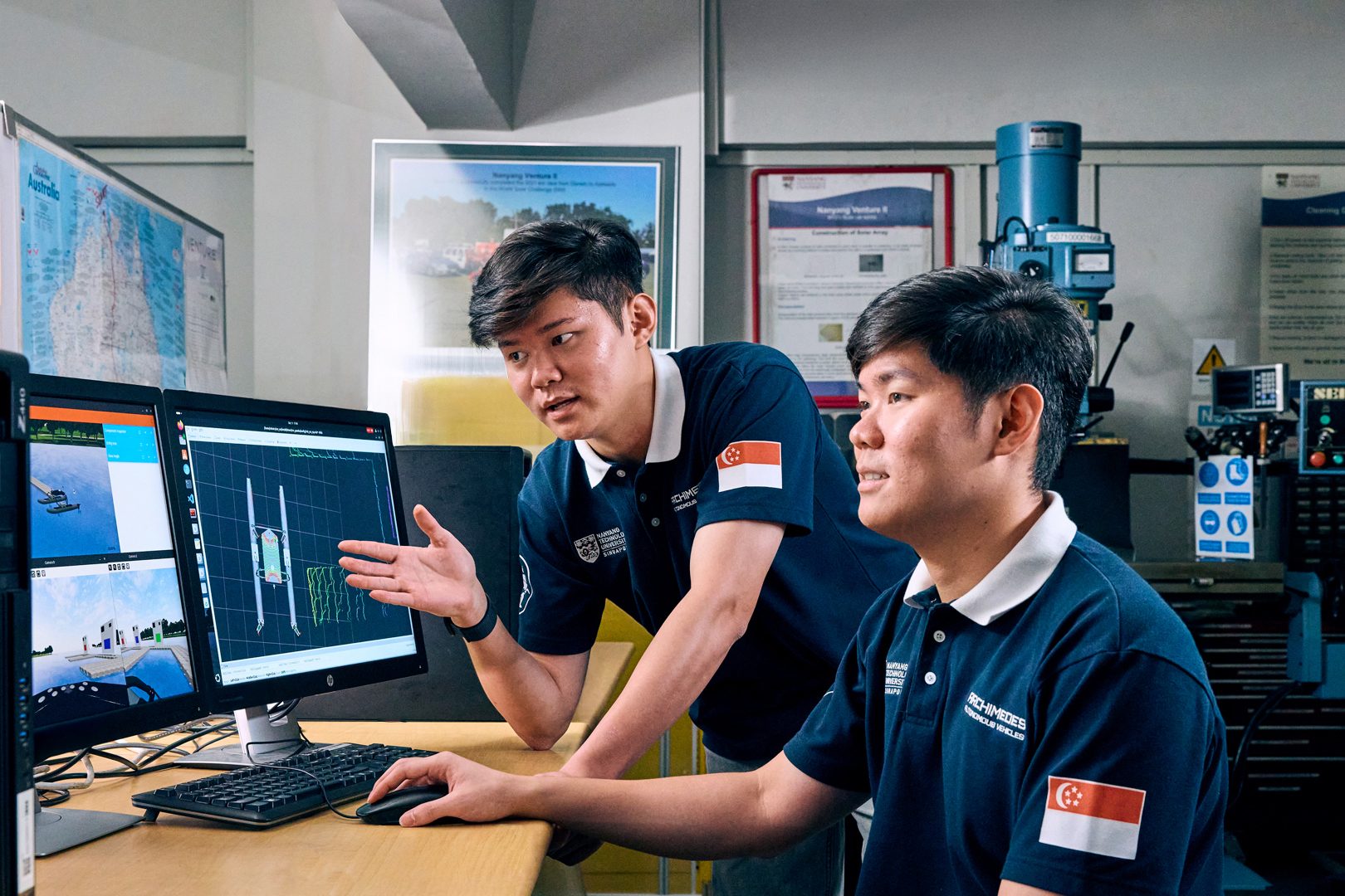 Two young men in Singapore university polos collaborate on technical designs on computer screens.