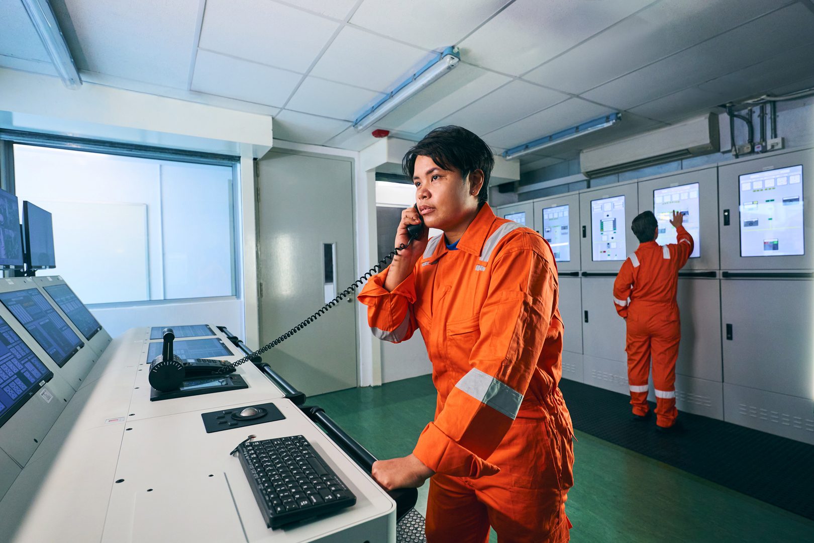 Two workers in orange coveralls in a control room, one on phone, one at control panels.