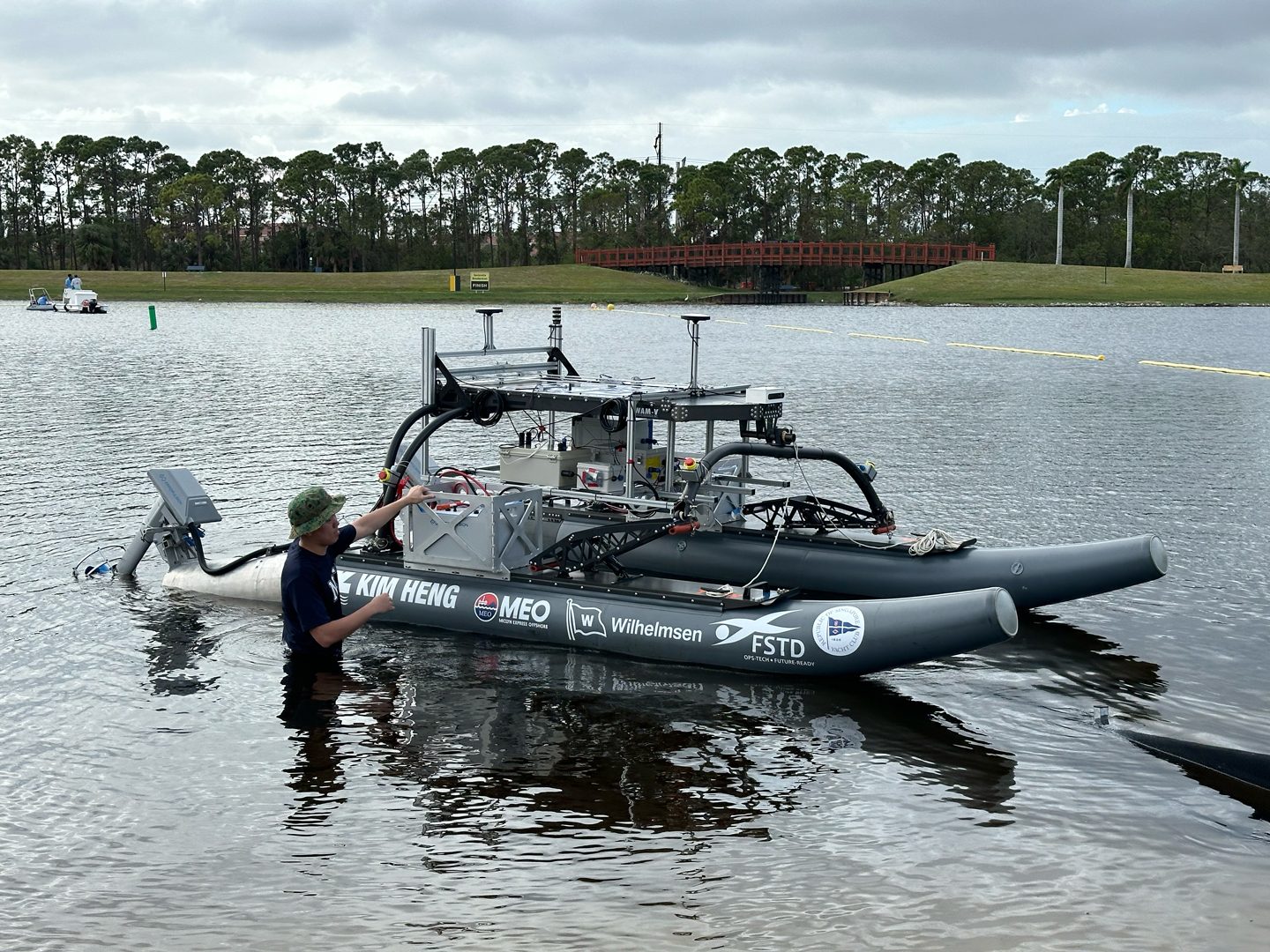 Man in water next to a grey catamaran-style autonomous boat. Lake, trees, and bridge in background.