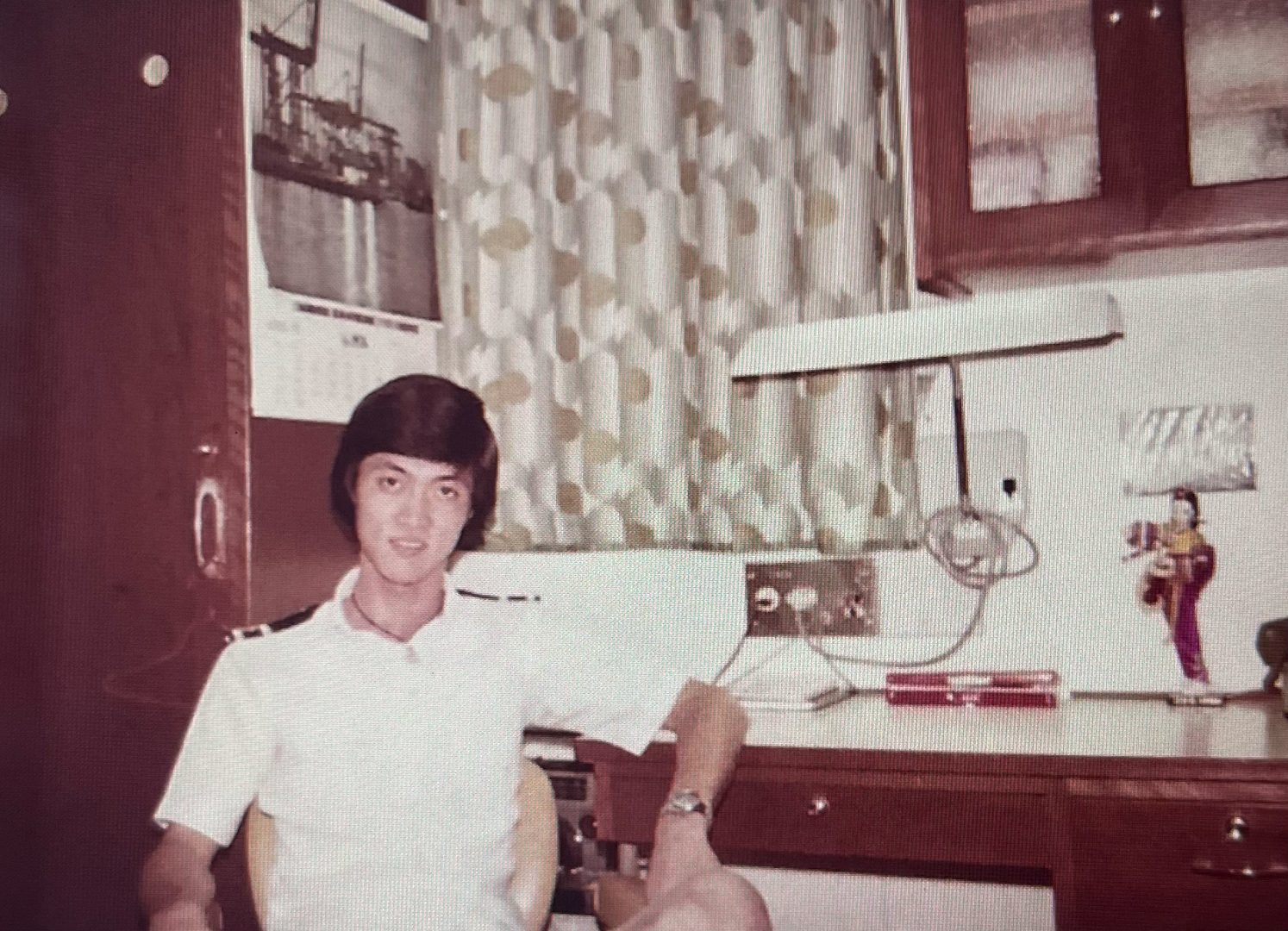 Young Asian man with a 70s hairstyle in a white polo shirt smiles while sitting at a desk.