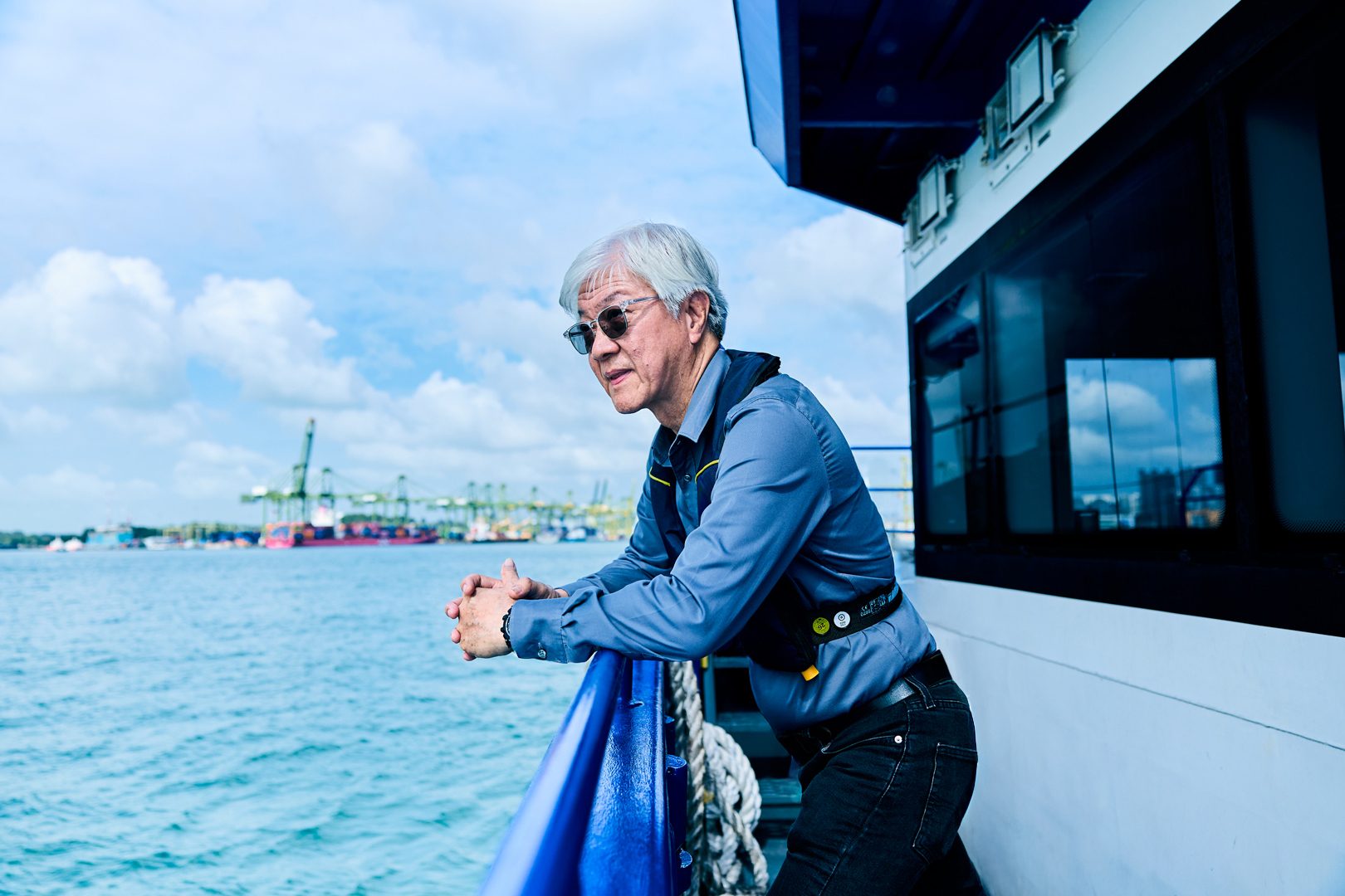 Man in sunglasses and life vest on boat, looking at a busy harbor with cranes.