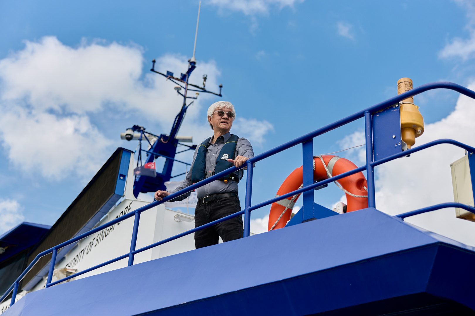 Man in a life vest and sunglasses on a boat deck, looking out at the cloudy sky.