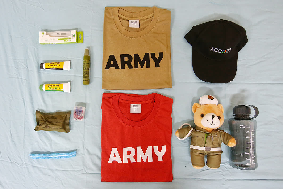 A flatlay of some items inside the women's assault packs, including (clockwise from top left) an oral thermometer, t-shirts, a cap, water bottle, a teddy bear, hair net, first aid dressing, ear buds and face paint.