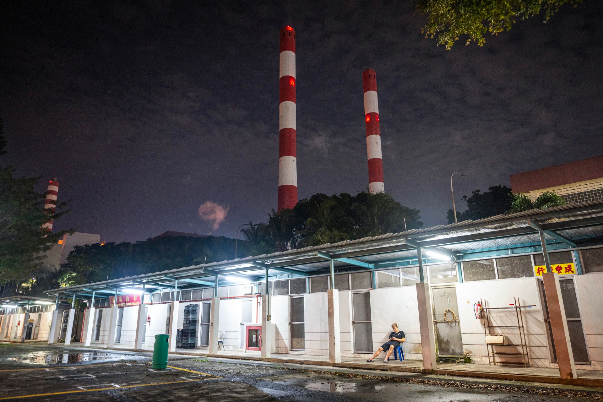 A man sitting outside a row of cold rooms, where fish merchants store their frozen seafood. The Senoko incineration plant can be seen in the background.