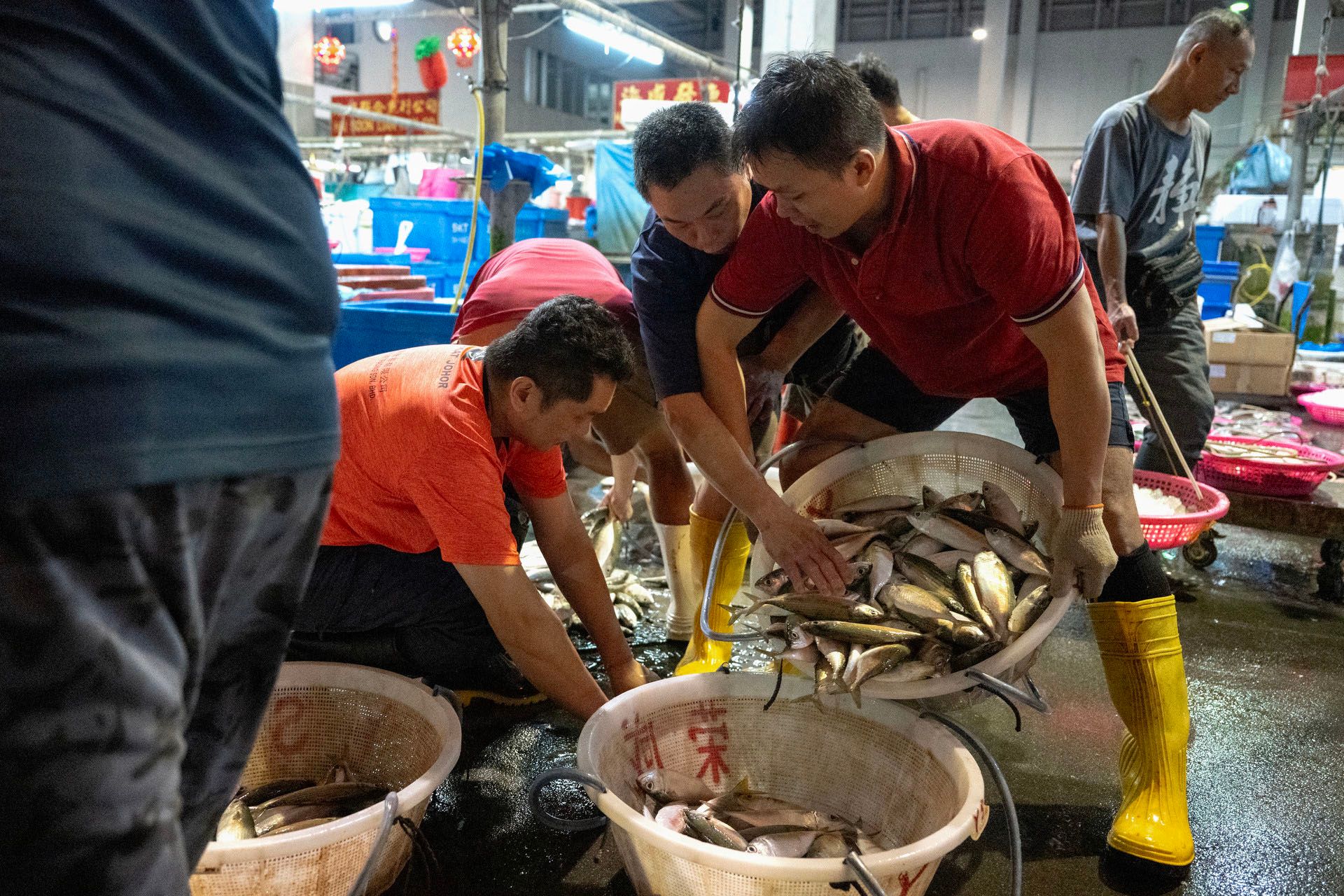 A fish seller playfully sneaking behind another seller to pick up a fish from the basket.