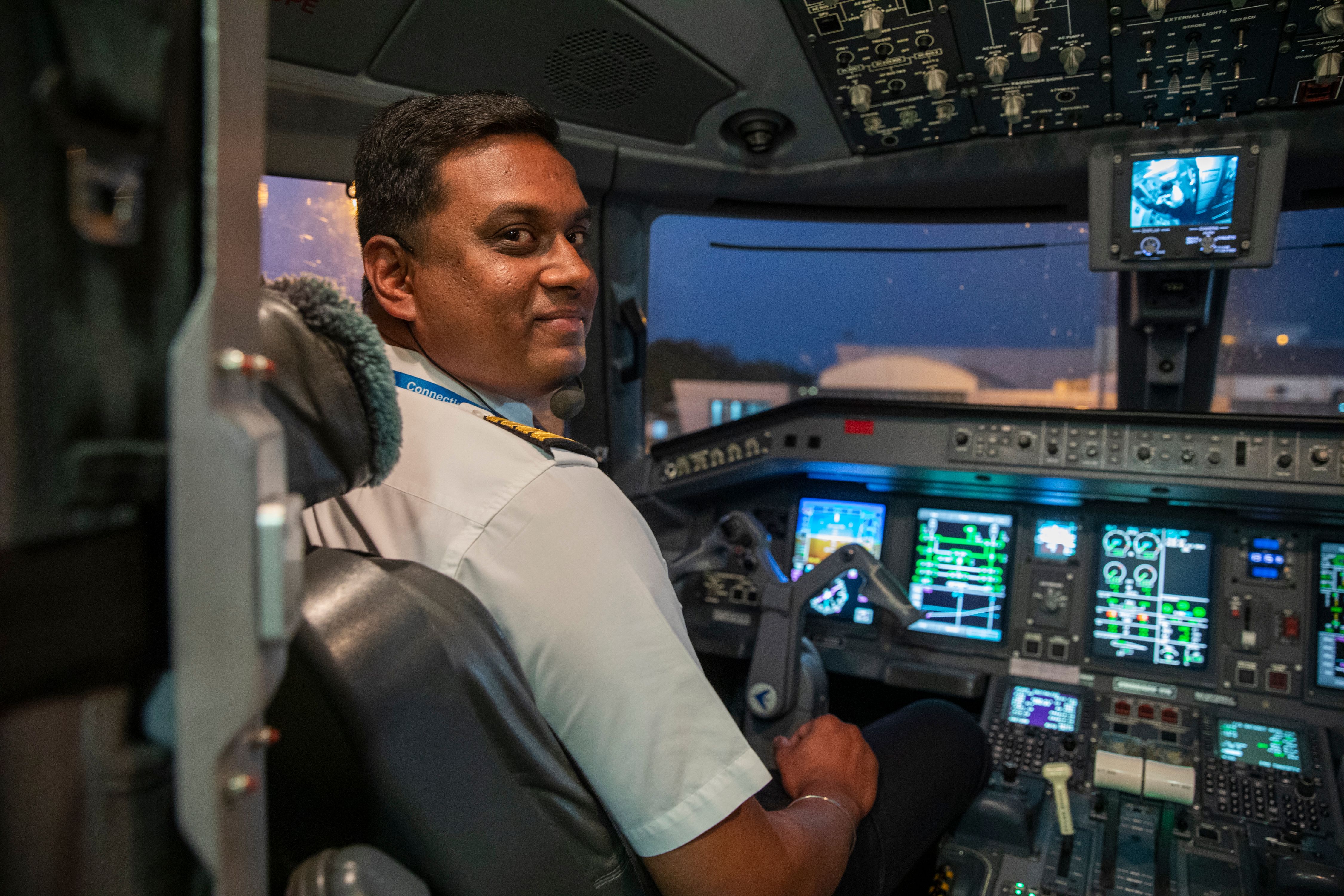 A uniformed male pilot in a cockpit smiles at the camera after landing in the evening in the Bengaluru airport.