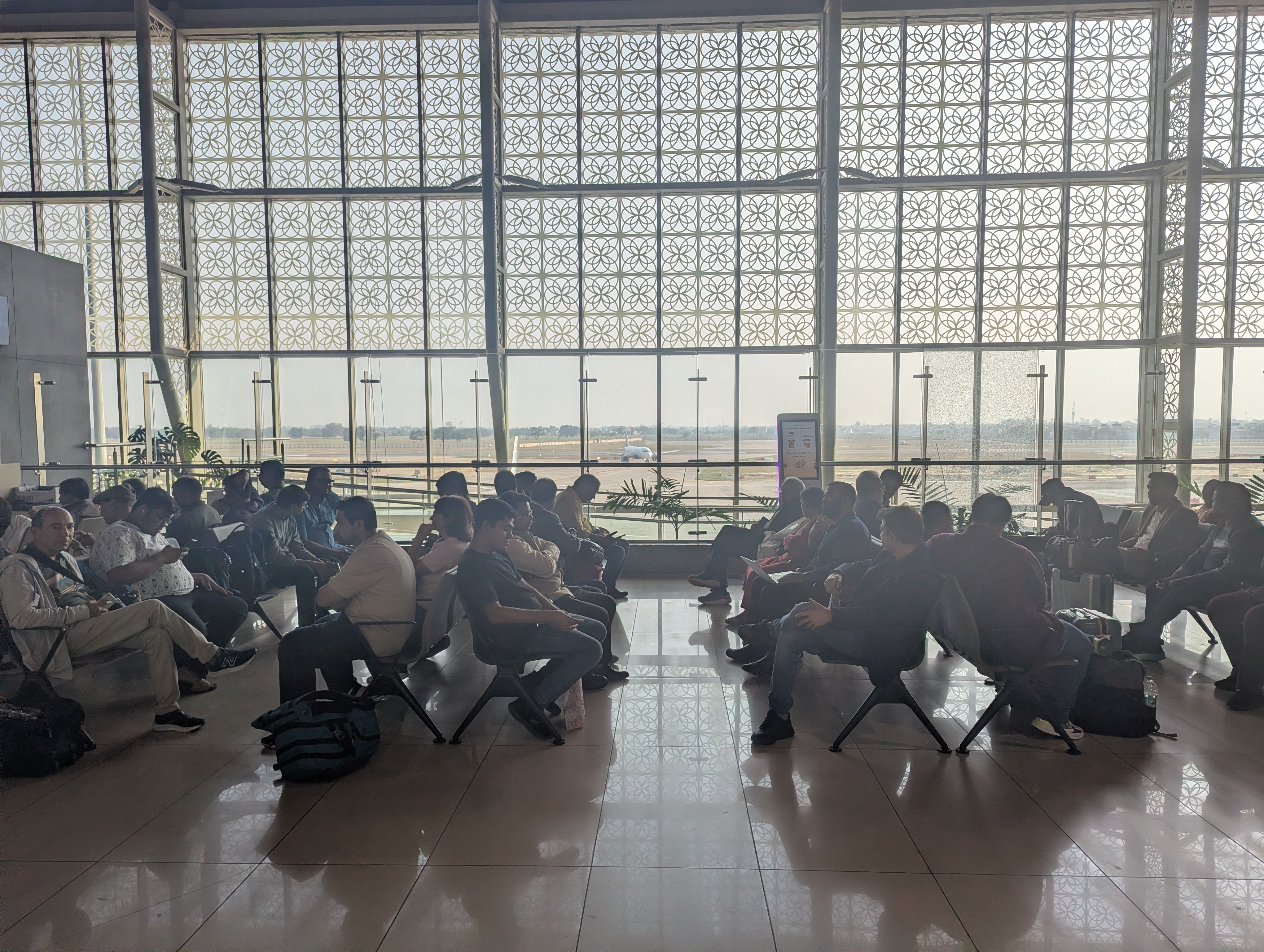Dozens of passengers sit on chairs, waiting at a boarding gate of the Lucknow airport, with sunlight pouring in from the glass walls, beyond which you can see an airplane taxiing on the runway.