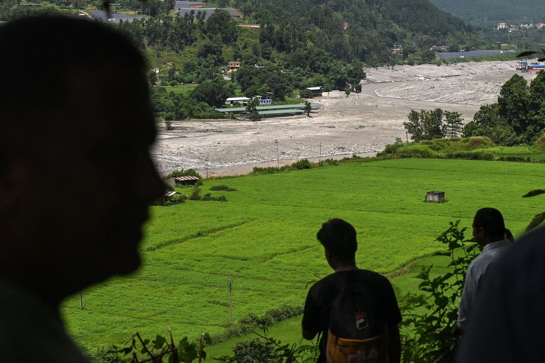 People shelter on higher ground and look across green fields towards a mud-filled section of the Trishuli River.