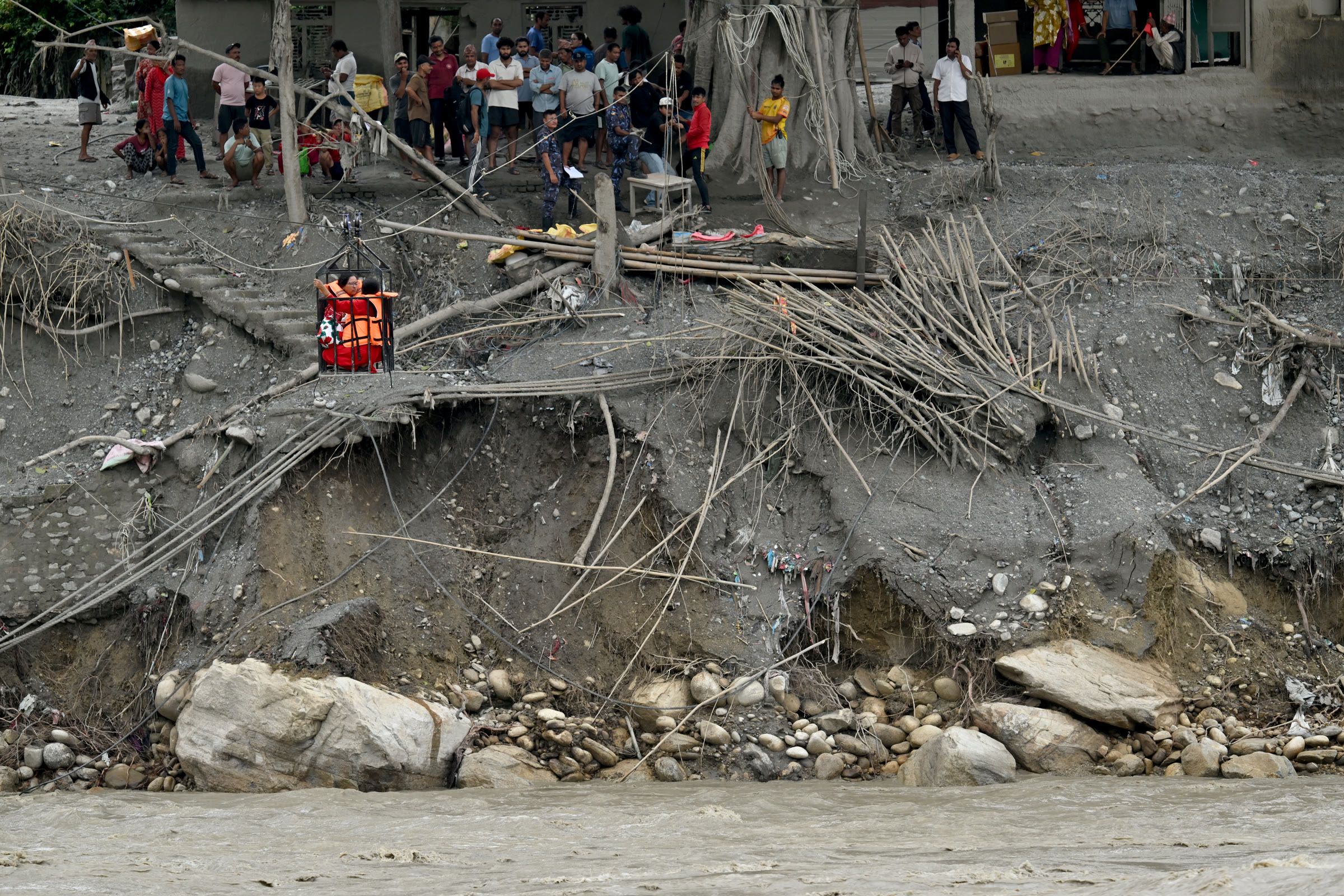 A resident in a life jacket crossing the Trishuli River in a metal cage as people watch from the eroded bank.