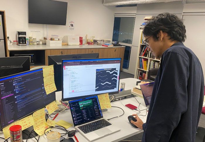 A photo of intern Azhari working on the creative coding to visualise psychoacoustics data on two computer monitors, surrounded by Post-its with scribbles on them.